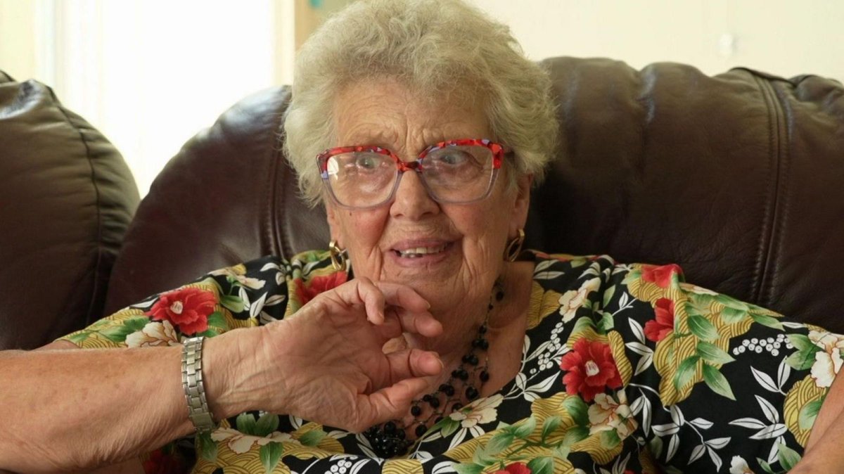 A woman in a floral top is sitting on a brown leather sofa, she has red rimmed glasses and short white hair.