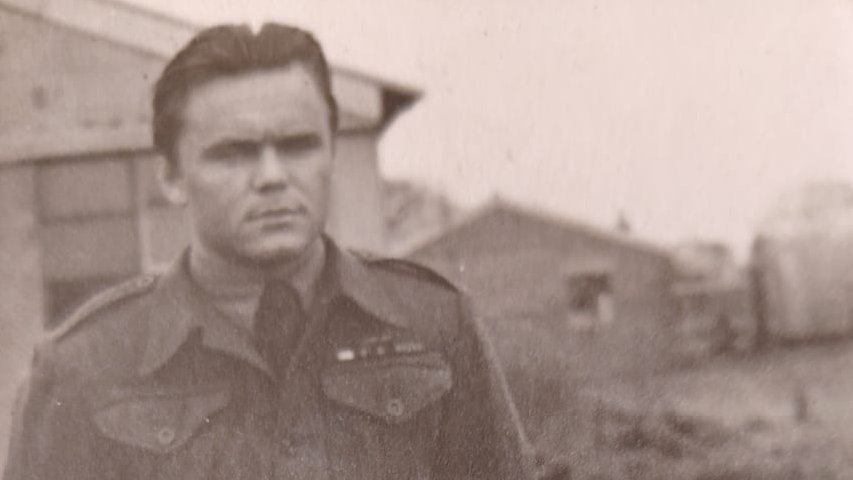 A black-and-white photograph shows a person in military uniform standing outdoors beside buildings, with open ground and additional structures visible in the background.
